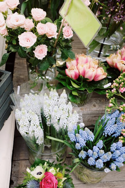 A variety of fresh floral arrangements displayed on a wooden surface, including a bouquet of pale pink roses with dark green leaves, a cluster of pink and cream tulips with lush green foliage, and sev