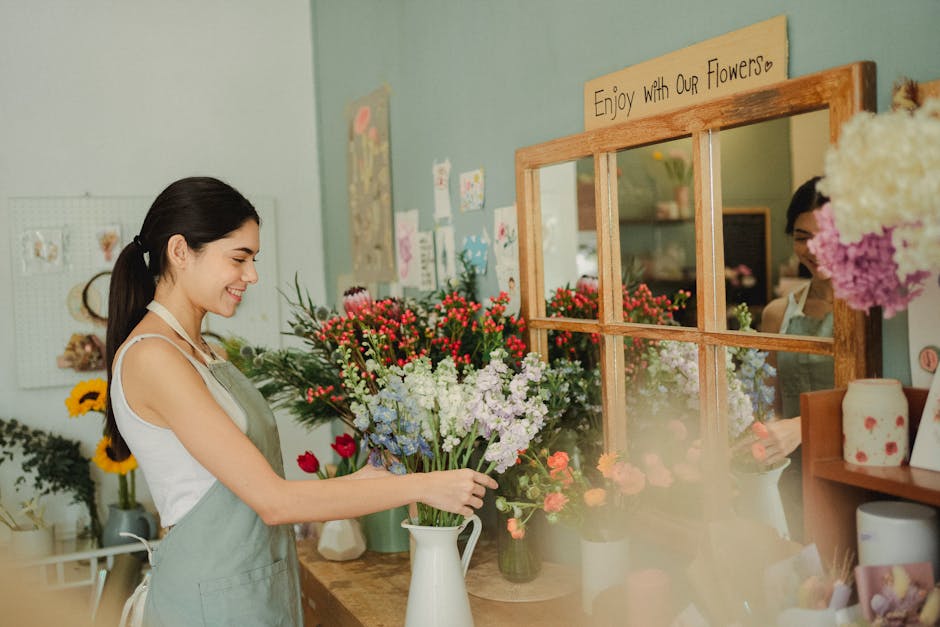 A young woman with dark hair tied back, smiling as she arranges a bouquet of freshly cut flowers in a white ceramic pitcher inside a flower shop. The bouquet features a mix of red, white, purple, and 
