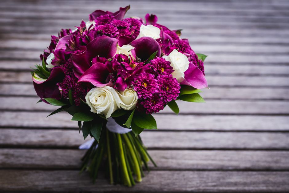 A beautifully arranged bouquet of fresh flowers resting on a wooden surface, featuring deep magenta calla lilies, vibrant pink dahlias, and purple orchids, complemented by white roses and lush green f
