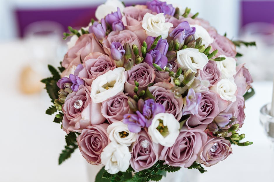 A floral bouquet arranged with soft pink roses, white lisianthus, and purple freesia, complemented by lush green foliage. The flowers are fresh with delicate, slightly ruffled petals, and the bouquet