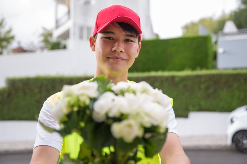 A delivery person wearing a red cap and a high-visibility vest holds out a bouquet of fresh white roses and green foliage towards the camera. The bouquet is wrapped in clear cellophane and placed in t